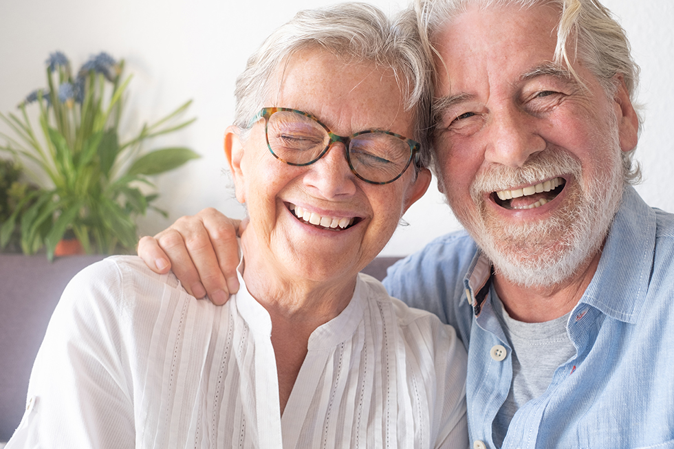 Elderly couple smiling warmly, sitting close with arms around each other on a couch.